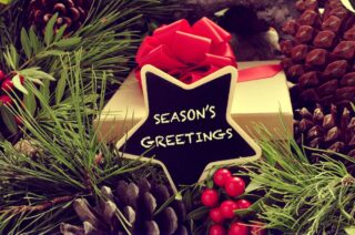 a star-shaped signboard with the text seasons greetings written in it, with a gift and some natural ornaments such pine cones and red berries in the background